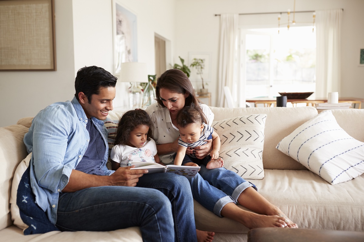 Hispanic couple sitting on the sofa reading a book at home with their baby son and young daughter
